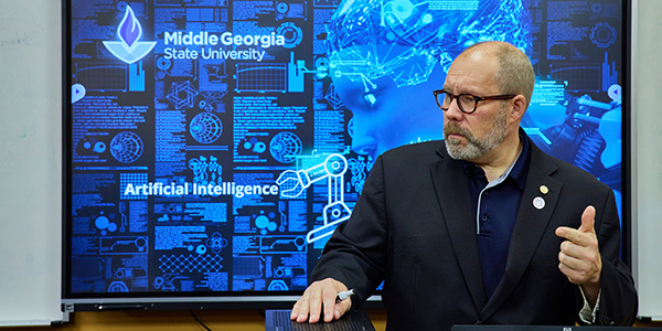 A man in a suit standing in front of a digital display showing artificial intelligence graphics and the Middle Georgia State University logo.